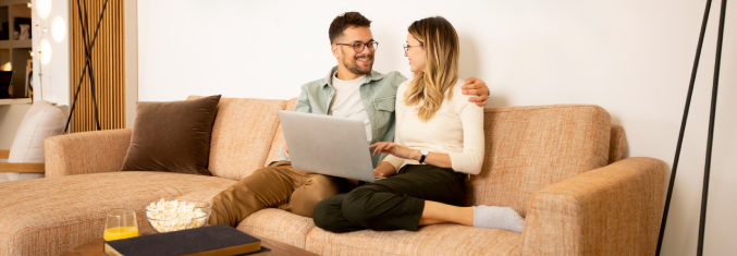 A couple sitting on a couch sharing a laptop, looking at eachother and discussing their finances